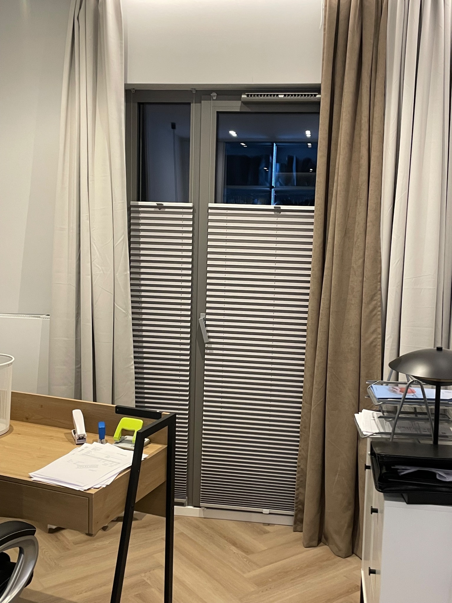 Zamknięte, poziome rolety plisowane w oknie, z widocznymi fałdami materiału, zasłonięte częściowo przez beżowe i białe zasłony. W tle widoczne biuro.