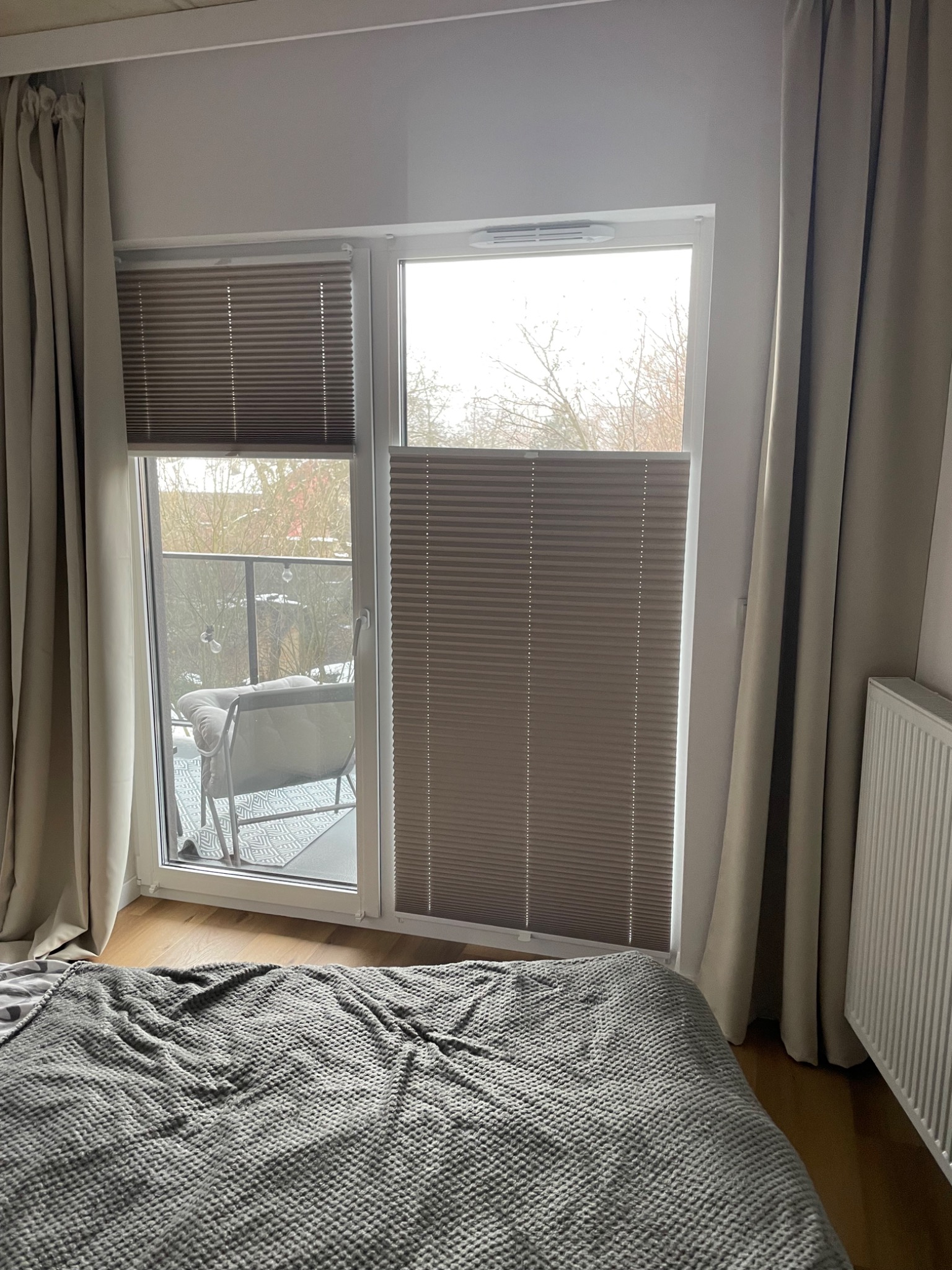 Beżowe rolety plisowane zamontowane w oknie balkonowym, widok z sypialni na balkon z krzesłem i lampkami. Częściowo zasłonięte beżowe zasłony po bokach okna.