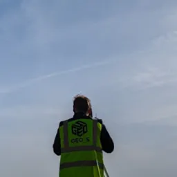 Mężczyzna w kamizelce odblaskowej z logo GEO_S obsługuje niwelator na trójnogu na polu, widoczny ślad samolotu na błękitnym niebie.