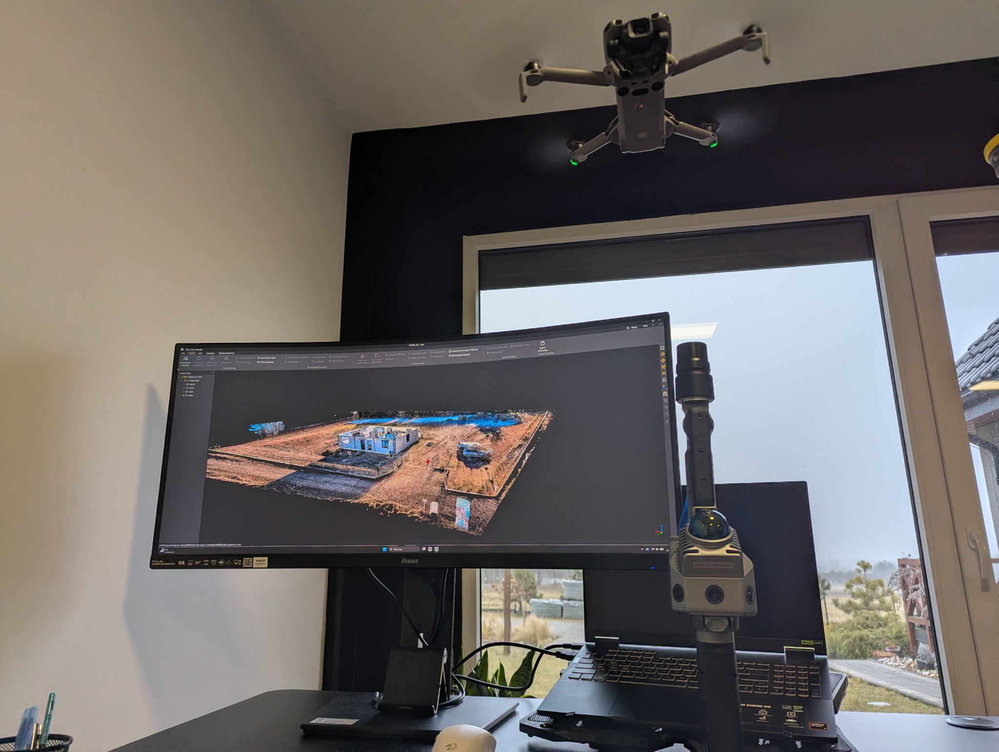 Dron w biurze nad stanowiskiem z monitorem prezentującym trójwymiarowy model budynku w trakcie budowy, obok skaner 3D. Usługi pomiarowe z powietrza.