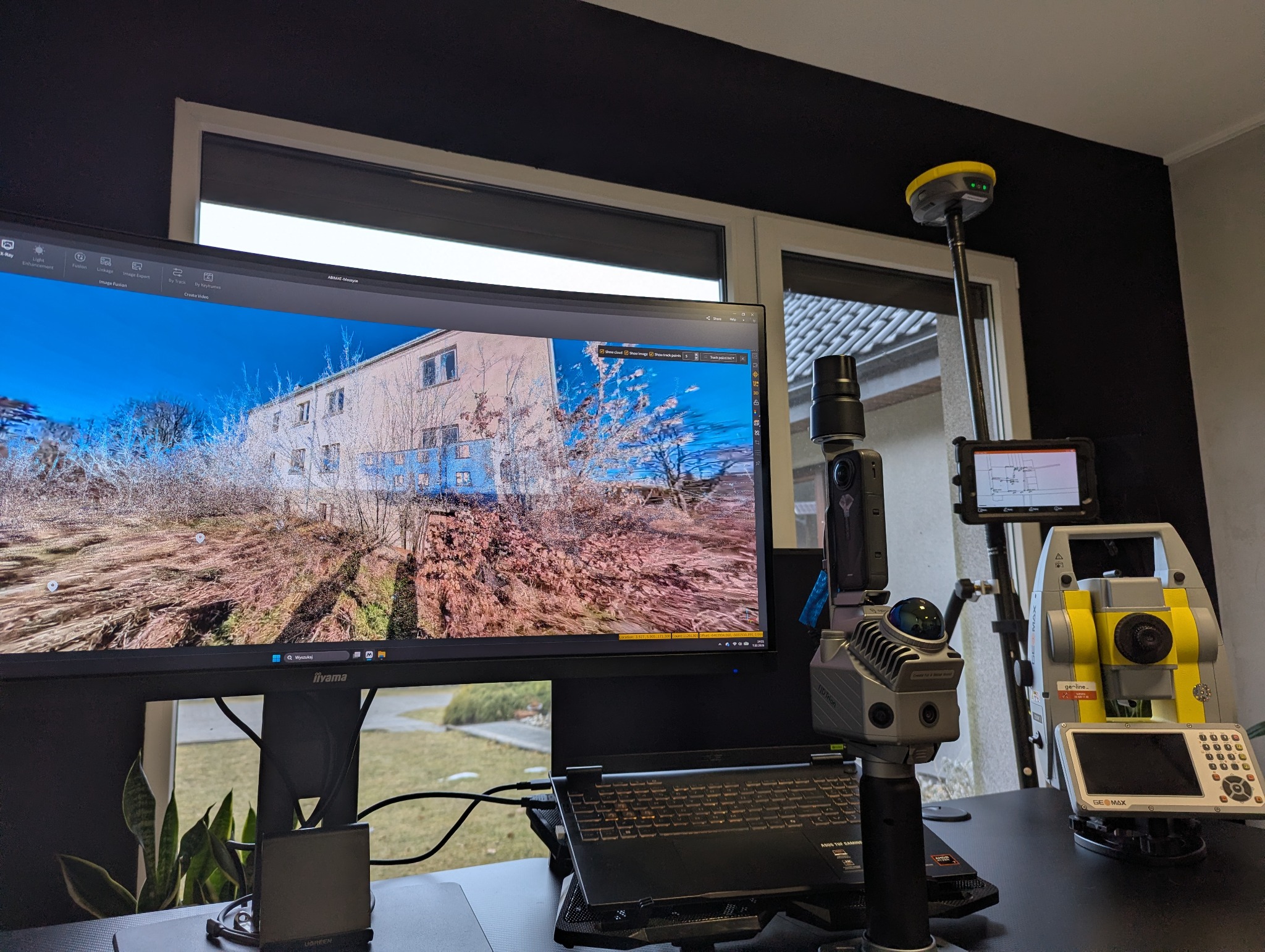 Stanowisko pracy z monitorem prezentującym model 3D budynku, obok sprzęt pomiarowy: tachimetr i skaner 3D. Widoczne okno w tle. Precyzyjne pomiary w budownictwie.