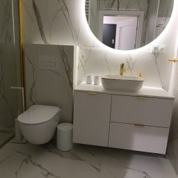Nowoczesna łazienka z marmurowymi płytkami, owalnym lustrem z podświetleniem LED, białą szafką z umywalką i złotymi akcentami. Widoczny fragment toalety i kosz.