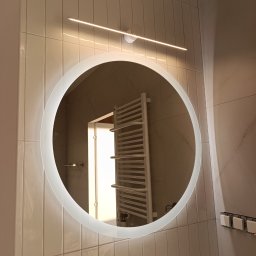 KOMFORT - Okrągłe lustro z podświetleniem LED w nowoczesnej łazience z beżowymi, pionowo ułożonymi płytkami. W odbiciu widoczny grzejnik drabinkowy.