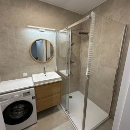 Nowoczesna łazienka w beżach: kabina prysznicowa z deszczownicą, umywalka z drewnianą szafką, okrągłe lustro i pralka. Minimalistyczny design, neutralna kolorystyka.