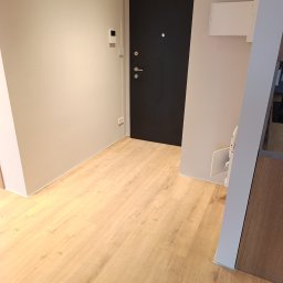 Jasne panele podłogowe w przedpokoju z czarnymi drzwiami wejściowymi i beżowymi ścianami. Widoczny fragment szafki z drewnianym frontem.