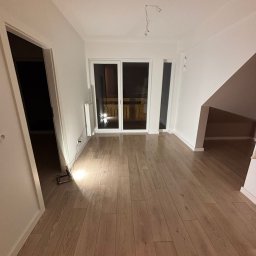 Jasne panele podłogowe w nowym, przestronnym pokoju z białymi ścianami i dużym oknem balkonowym. Widoczne niedokończone prace elektryczne na suficie.