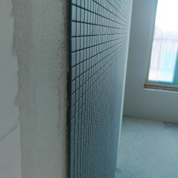 KOMFORT - Ściana wyłożona mozaiką w odcieniach szarości, widoczna z bliska pod kątem. Fragment ściany obok w stanie surowym, okno w tle. Minimalistyczny kadr, ostre światło.