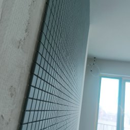 KOMFORT - Narożnik ściany z surowym tynkiem i fragmentem ściany wyłożonej mozaiką w odcieniach szarości. W tle widoczne okno i fragment sufitu z lampą.