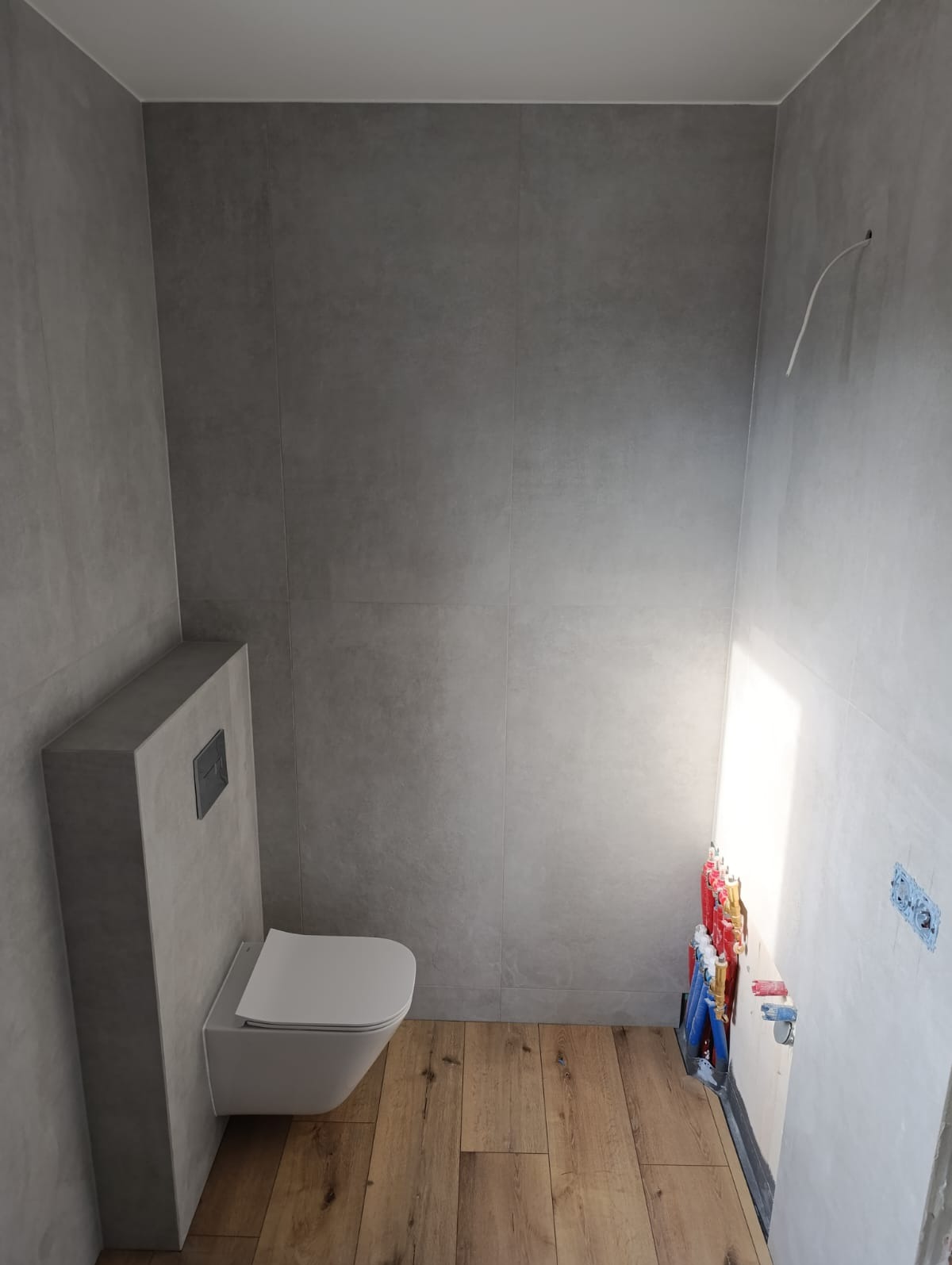 Nowoczesna łazienka w trakcie prac wykończeniowych: podwieszana toaleta na tle szarych płytek, drewnopodobne panele podłogowe, widoczne instalacje wodne.