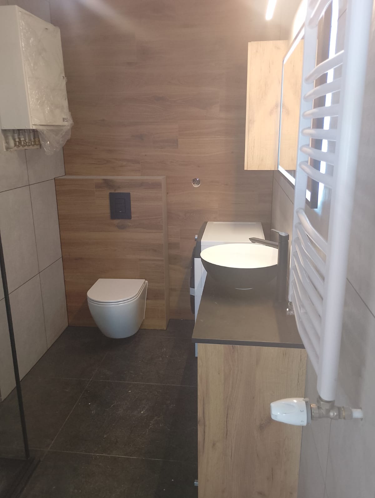 Nowoczesna łazienka z wiszącą toaletą, umywalką nablatową w kolorze czarnym, grzejnikiem drabinkowym i płytkami imitującymi drewno na ścianach.