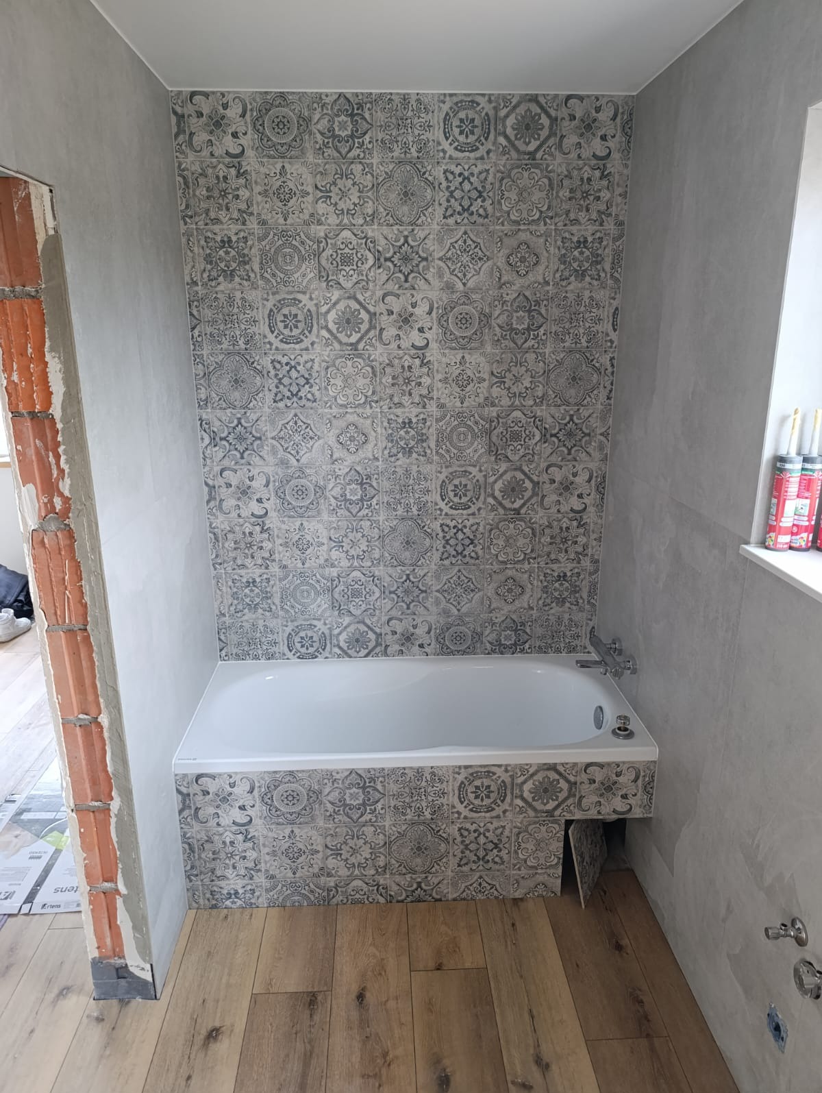 Wanna obudowana płytkami z wzorem patchworkowym, dopasowanymi do płytek na ścianie; widoczny fragment niedokończonej ściany z cegły i fragment podłogi z paneli imitujących drewno.