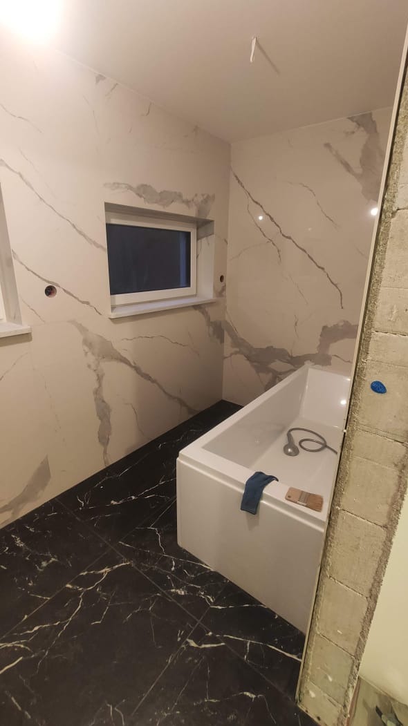 Nowoczesna łazienka w trakcie wykańczania: biała wanna, ściany wyłożone płytkami imitującymi marmur, czarna podłoga z płytek z białymi żyłami, widoczne niedokończone prace przy ścianie.