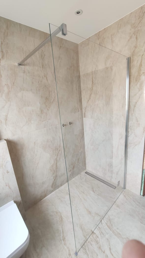 Kabina prysznicowa walk-in z transparentnego szkła, zamontowana w rogu łazienki wyłożonej płytkami imitującymi marmur, widoczny odpływ liniowy w podłodze.