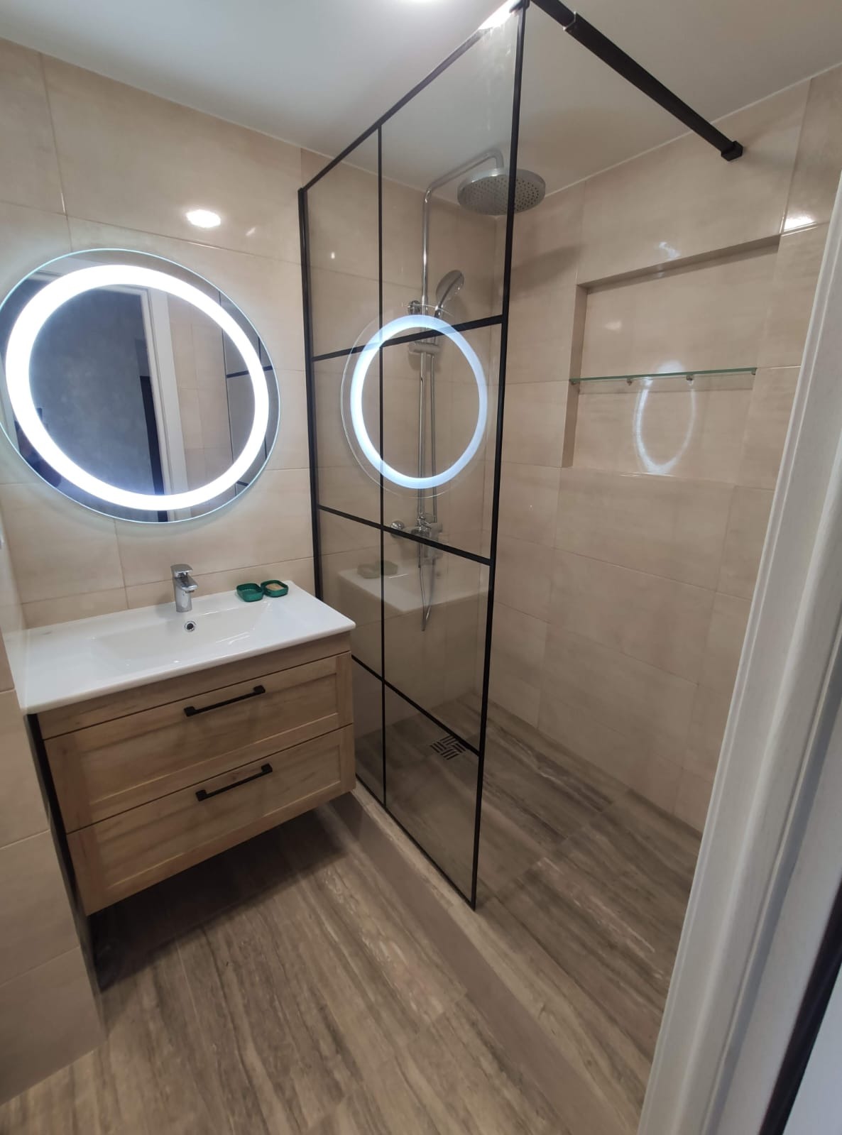 Nowoczesna łazienka z umywalką na szafce z drewnianymi frontami, okrągłym lustrem z podświetleniem LED i kabiną prysznicową typu walk-in z czarnymi profilami i deszczownicą.