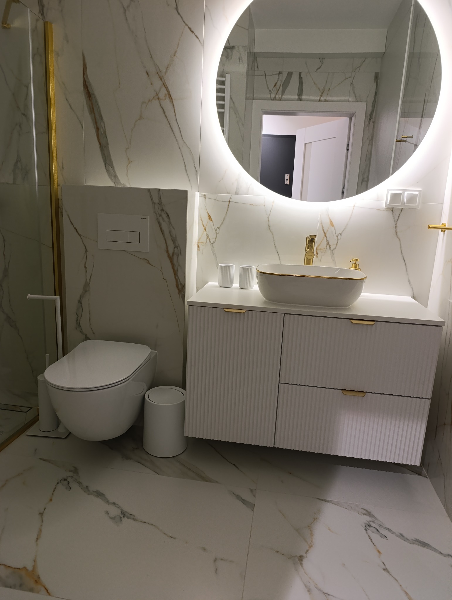 Nowoczesna łazienka z marmurowymi płytkami, owalnym lustrem z podświetleniem LED, białą szafką z umywalką i złotymi akcentami. Widoczny fragment toalety i kosz.