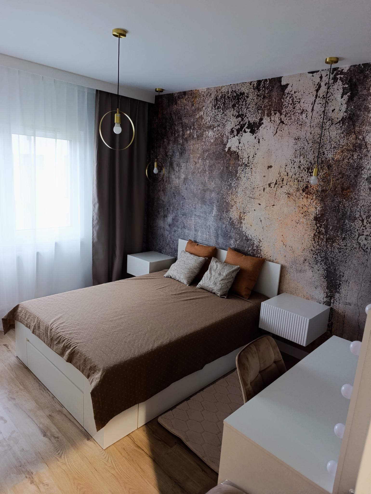 Sypialnia z łóżkiem, szafkami nocnymi i toaletką. Ściana z efektem betonu, złote lampy wiszące. Parkiet z jasnego drewna. Nowoczesny design wnętrza.