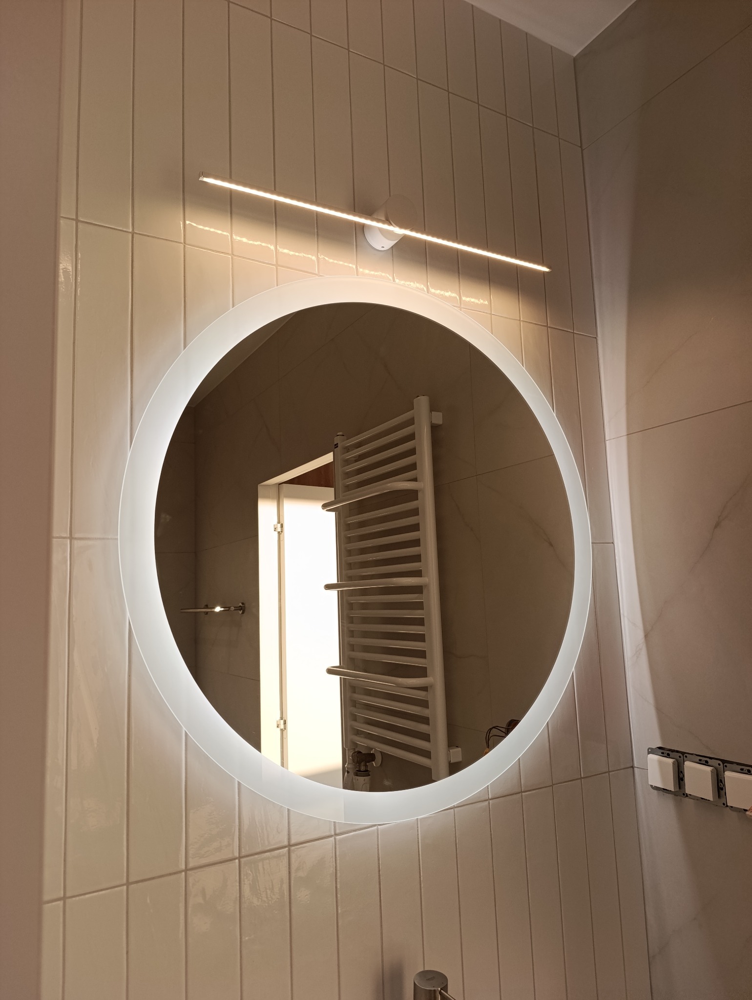 Okrągłe lustro z podświetleniem LED w nowoczesnej łazience z beżowymi, pionowo ułożonymi płytkami. W odbiciu widoczny grzejnik drabinkowy.
