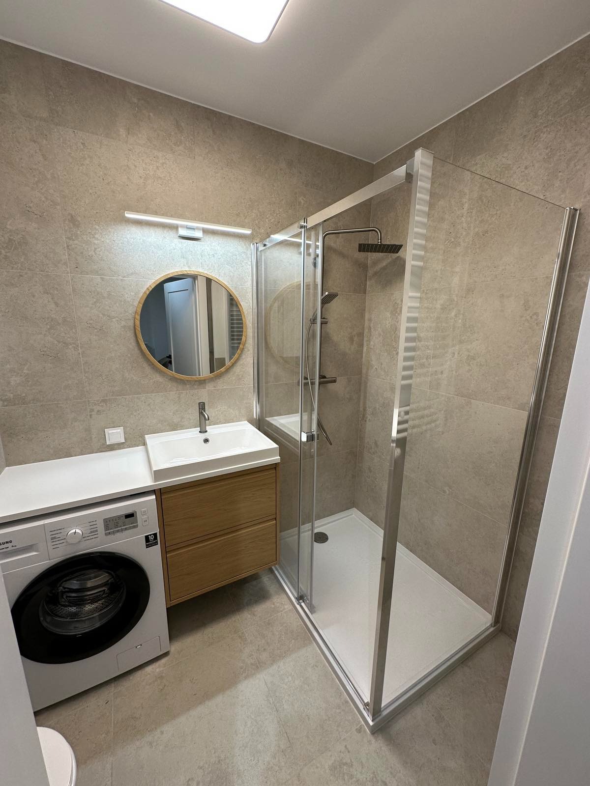 Nowoczesna łazienka w beżach: kabina prysznicowa z deszczownicą, umywalka z drewnianą szafką, okrągłe lustro i pralka. Minimalistyczny design, neutralna kolorystyka.