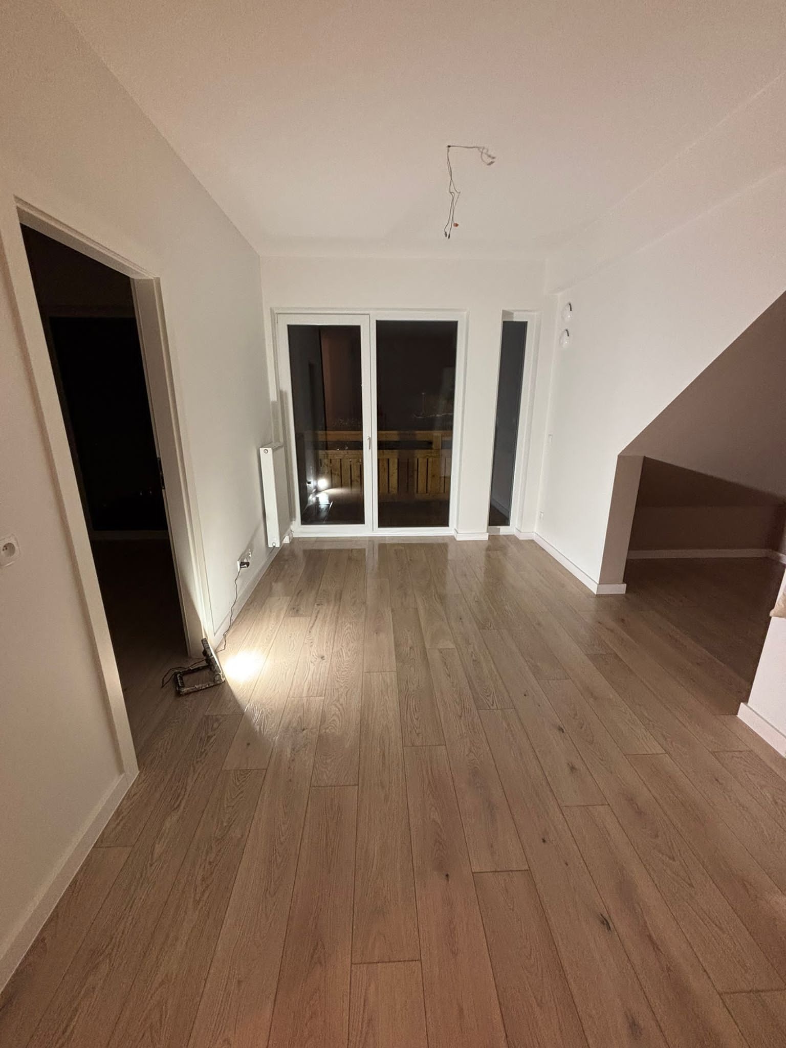 Jasne panele podłogowe w nowym, przestronnym pokoju z białymi ścianami i dużym oknem balkonowym. Widoczne niedokończone prace elektryczne na suficie.