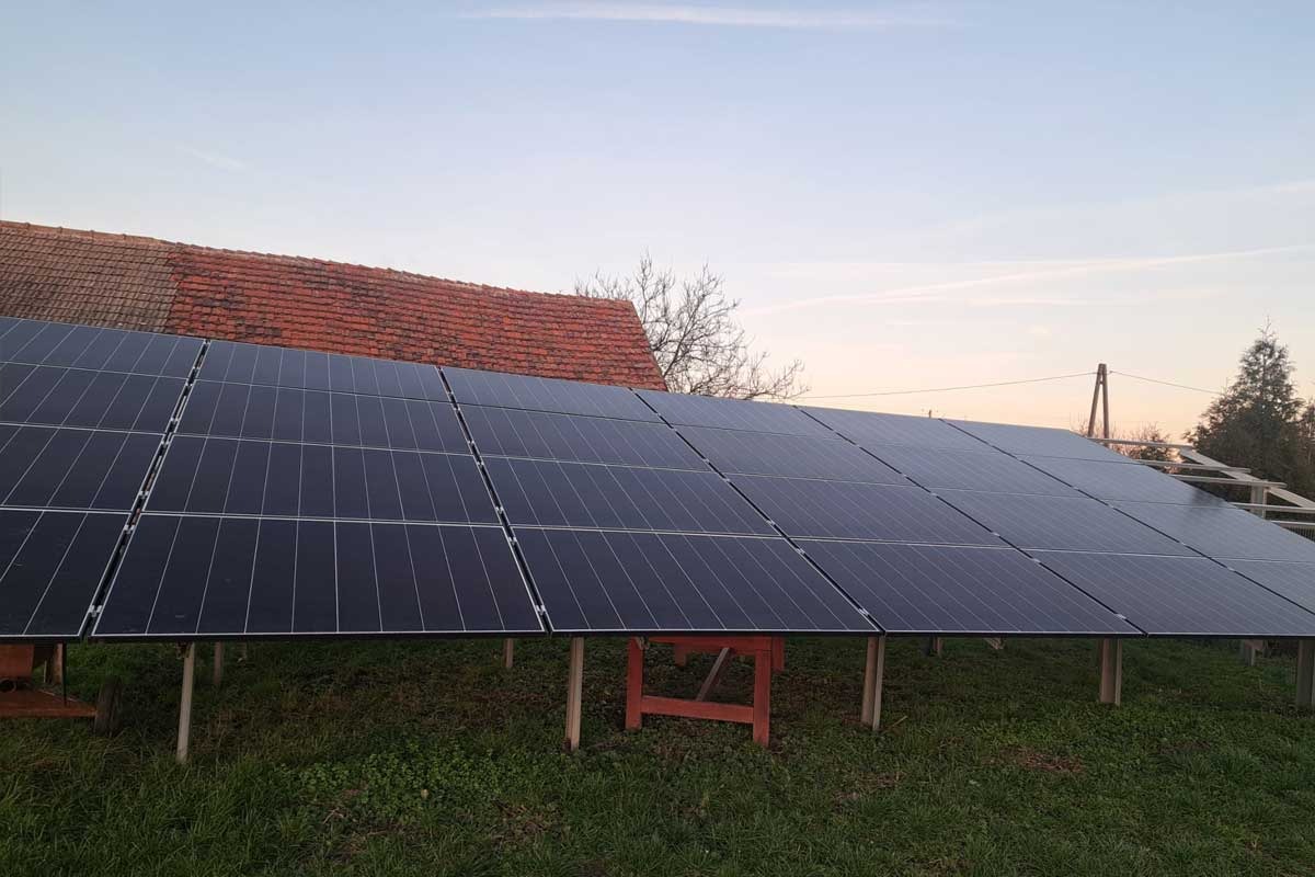 Naziemna instalacja fotowoltaiczna na drewnianej konstrukcji, widoczny dach domu w tle, pora dnia: wczesny ranek lub późne popołudnie.