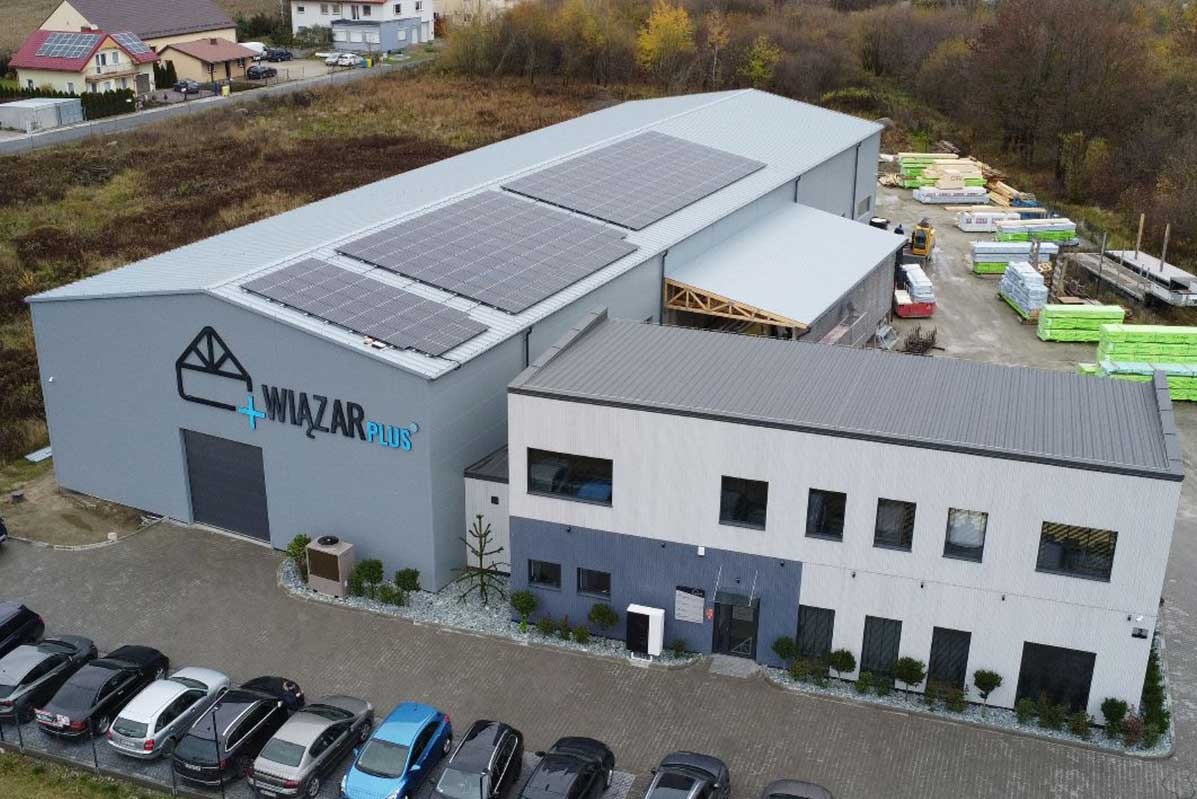 Widok z góry na duży budynek przemysłowy z panelami słonecznymi na dachu, zaparkowanymi samochodami i składowanym drewnem w tle.
