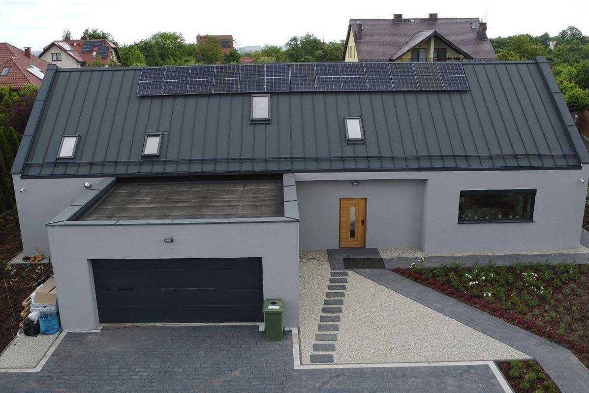 Nowoczesny dom jednorodzinny z szarą elewacją i panelami słonecznymi na grafitowym dachu, widok z góry na podjazd z kostki i mały ogródek.