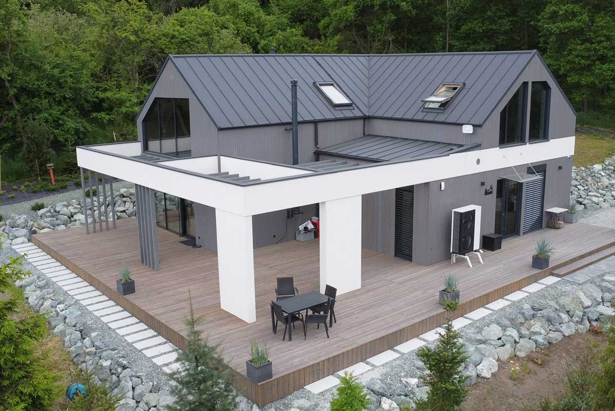 Nowoczesny dom jednorodzinny z szarą elewacją, grafitowym dachem i dużym drewnianym tarasem, na którym znajduje się czarny stół z krzesłami, widoczny system grzewczy na zewnątrz budynku...