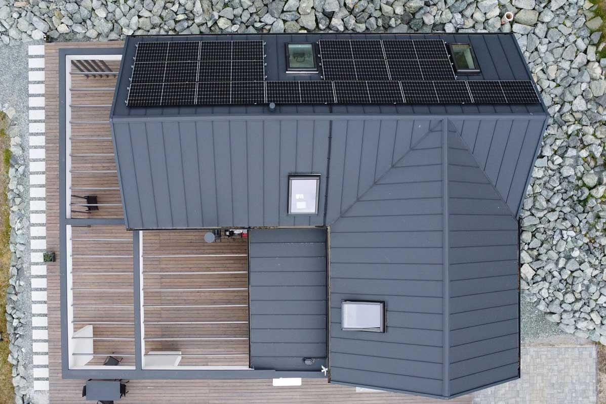 Widok z góry na dom z szarym dachem blaszanym, panelami słonecznymi i świetlikami, otoczony kamieniami i drewnianym tarasem z meblami.