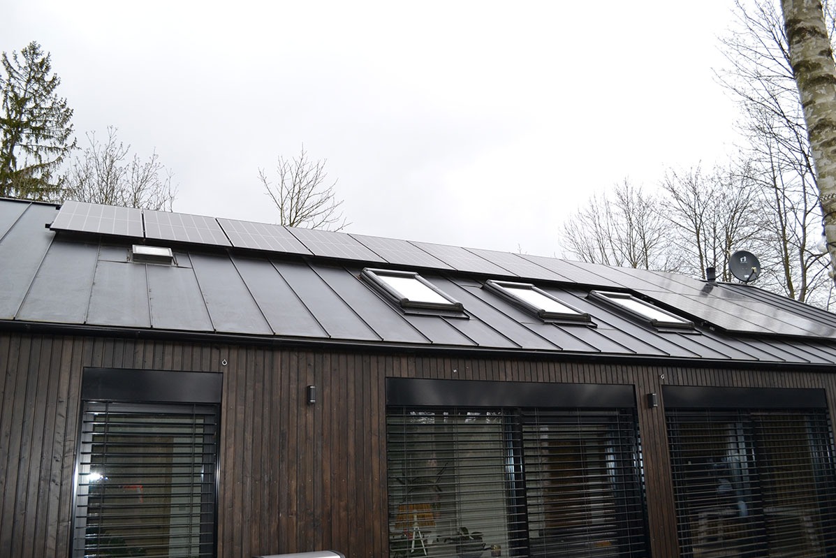 Ciemny, nowoczesny dom z panelami słonecznymi na dachu, trzema oknami dachowymi i zewnętrznymi roletami.