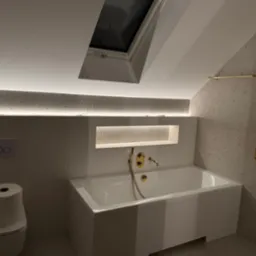 Biała łazienka na poddaszu z oknem dachowym, podświetlanym blatem z terazzo i złotą armaturą, widoczny sedes z papierem toaletowym.