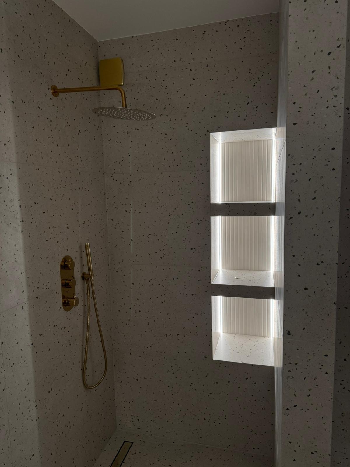 Nowoczesna kabina prysznicowa z armaturą w kolorze złotym i wnękami oświetlonymi LED, wykończona płytkami imitującymi lastryko.