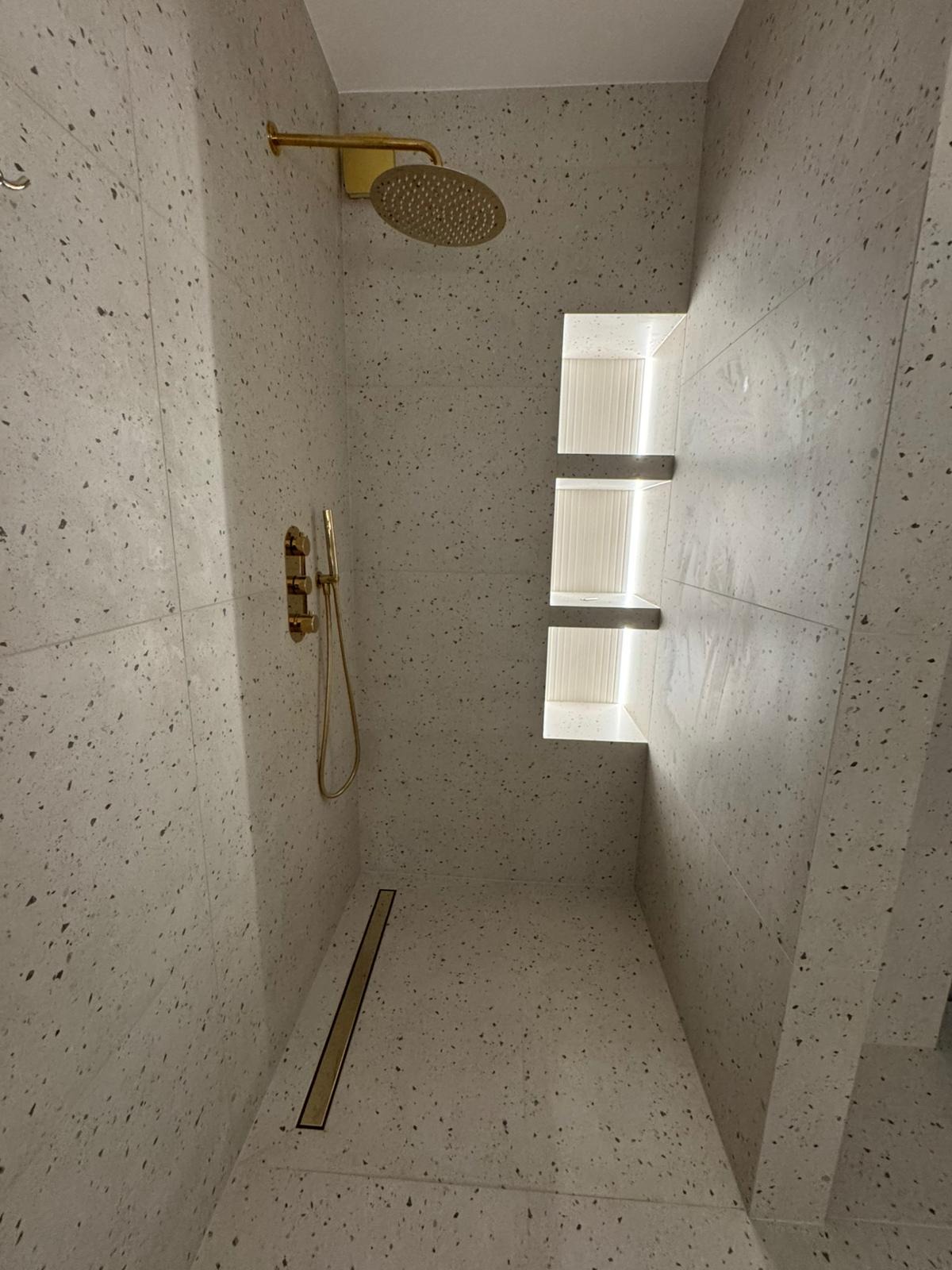 Nowoczesny prysznic walk-in z mozaikową okładziną ścian i podłogi, złotą armaturą i wnękami z podświetleniem LED.