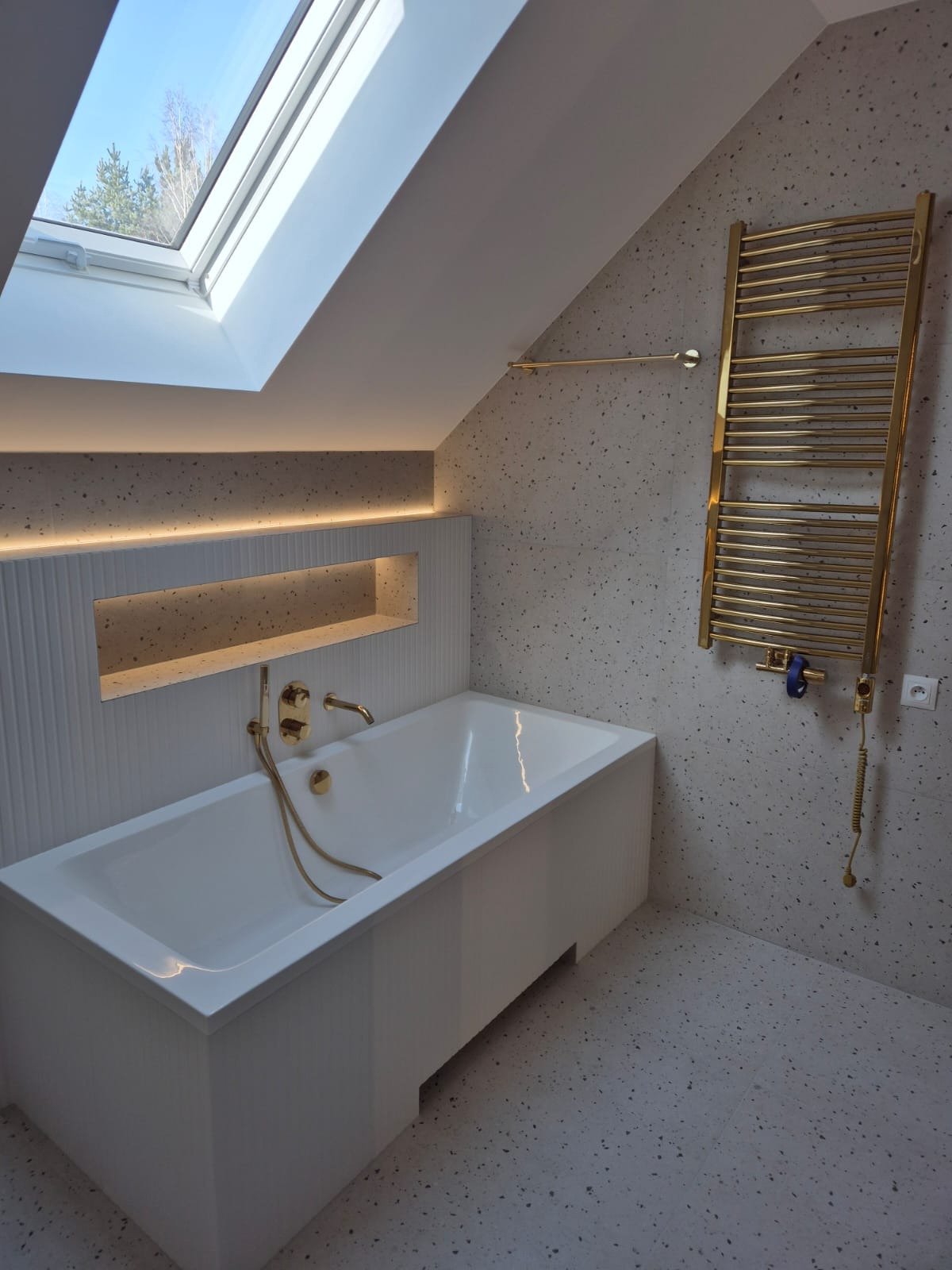 Nowoczesna łazienka na poddaszu z białą wanną, złotą armaturą i grzejnikiem, oświetleniem LED w niszy ściennej i oknem dachowym z widokiem na drzewa.