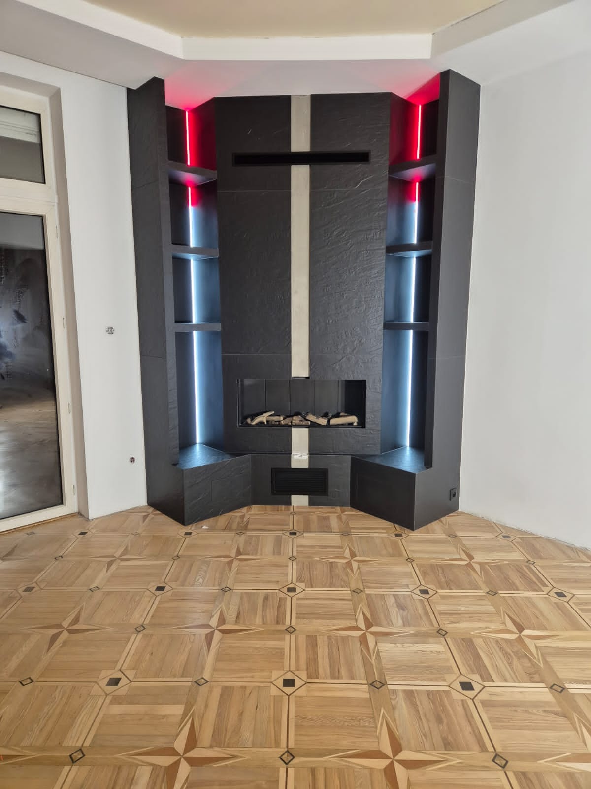 Nowoczesny kominek z czarnej płyty z wbudowanymi półkami oświetlonymi czerwonym i niebieskim światłem, na podłodze parkiet w geometryczny wzór.