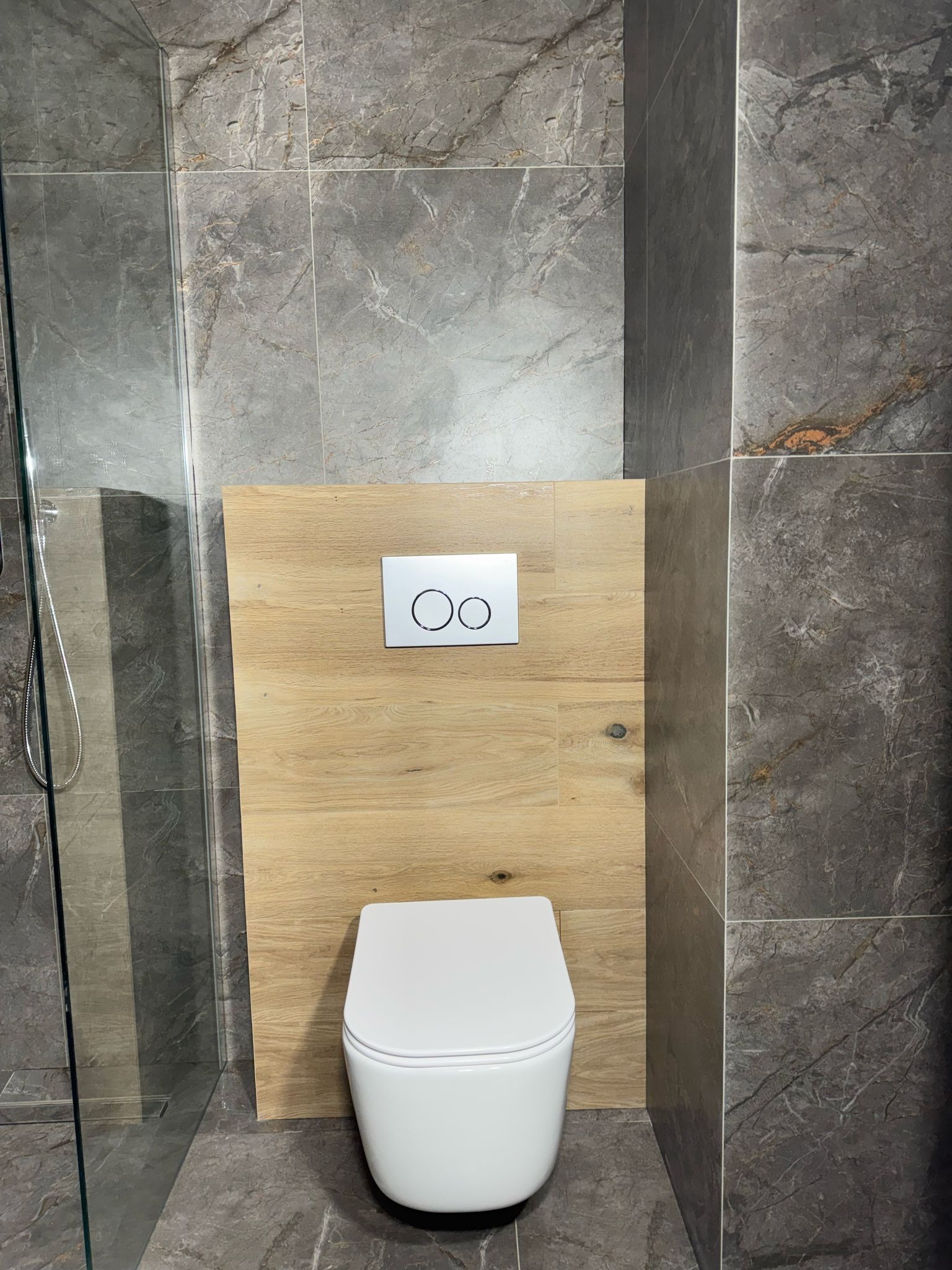 Nowoczesna łazienka z szarą marmurową płytką, drewnianą zabudową stelaża WC i szklaną kabiną prysznicową.