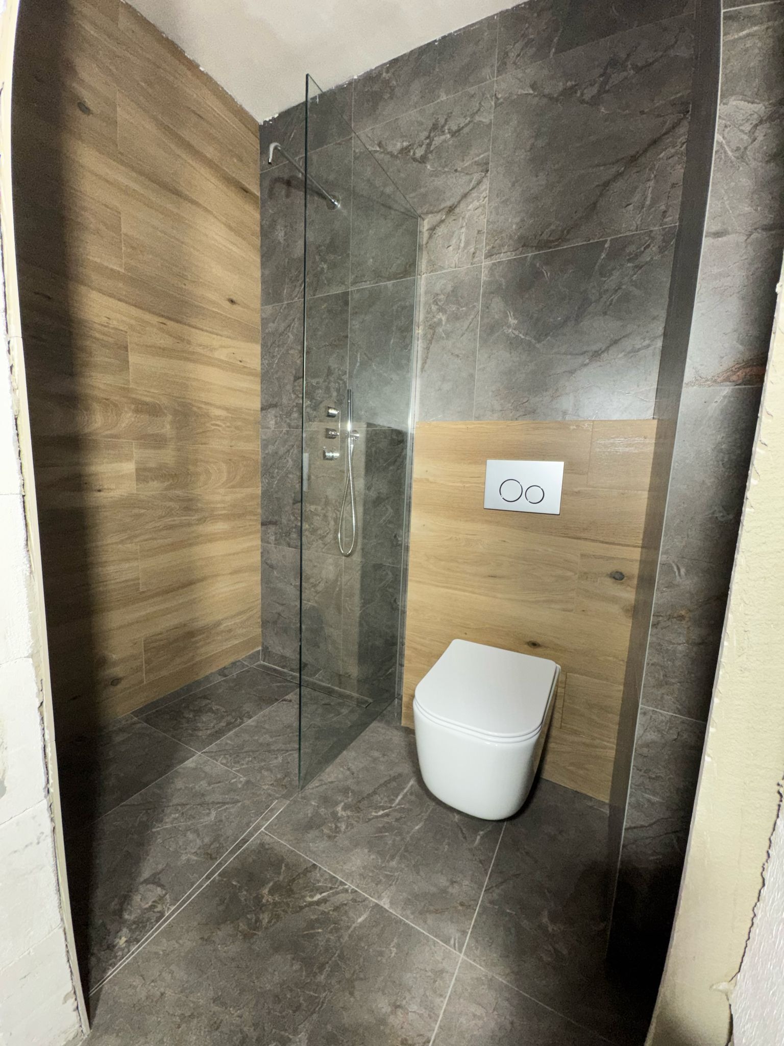 Nowoczesna łazienka w trakcie wykańczania: drewnopodobne i marmurowe płytki, szklana kabina prysznicowa z deszczownicą, podwieszana toaleta z chromowanym przyciskiem spłukującym.