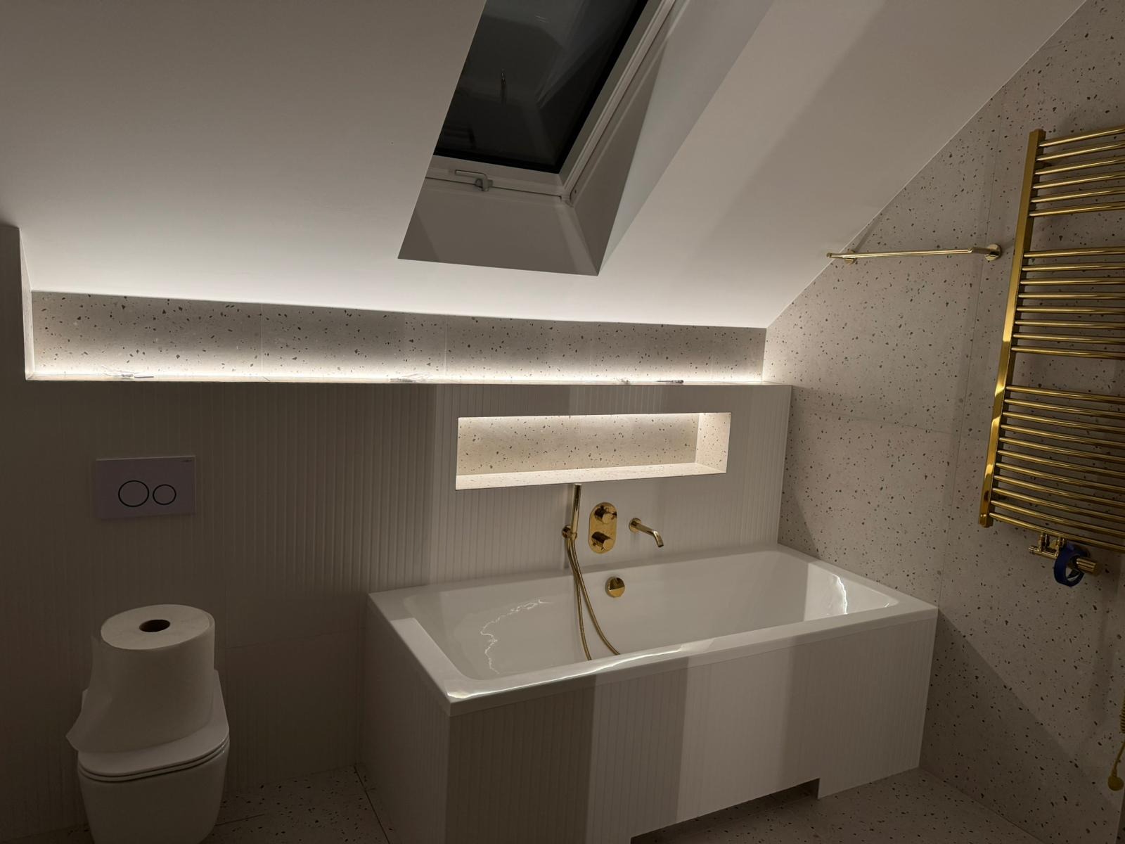 Biała łazienka na poddaszu z oknem dachowym, podświetlanym blatem z terazzo i złotą armaturą, widoczny sedes z papierem toaletowym.