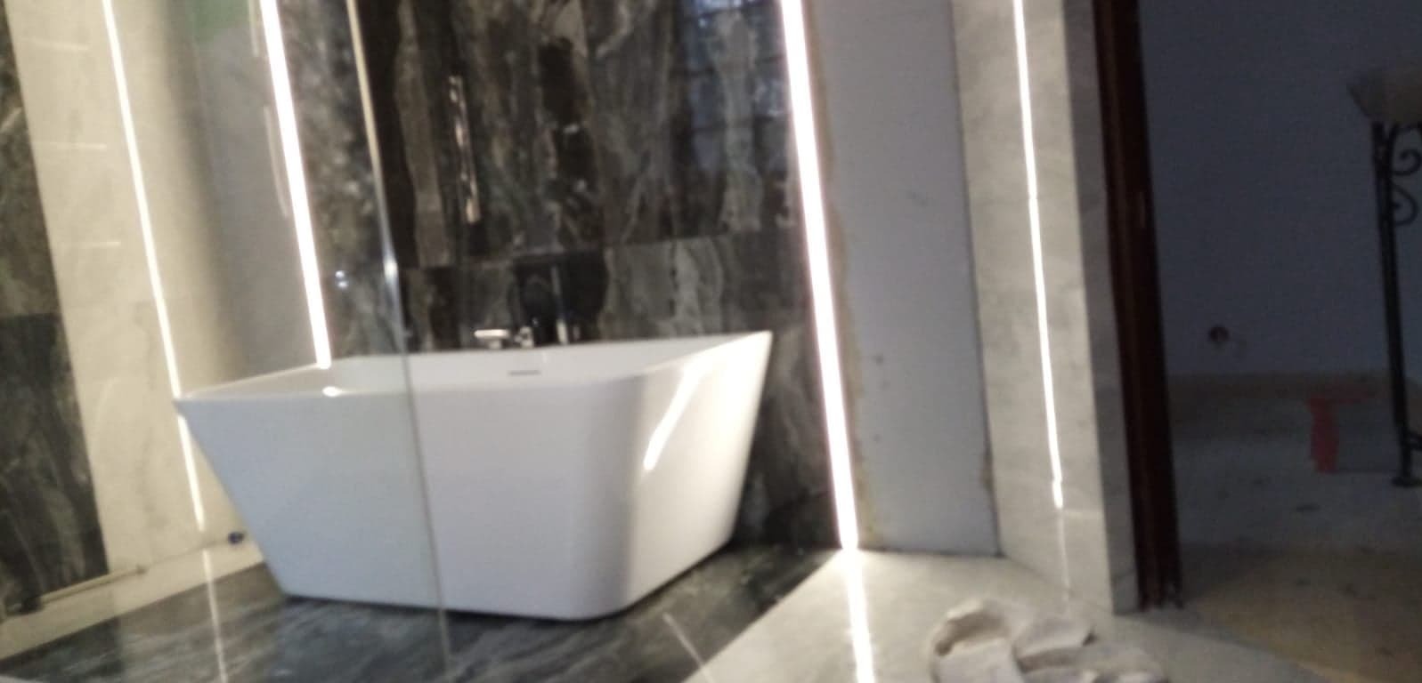 Nowoczesna łazienka z wolnostojącą wanną na tle ściany z szarego marmuru, oświetlona ledowym światłem. Widoczny fragment podłogi i drzwi.