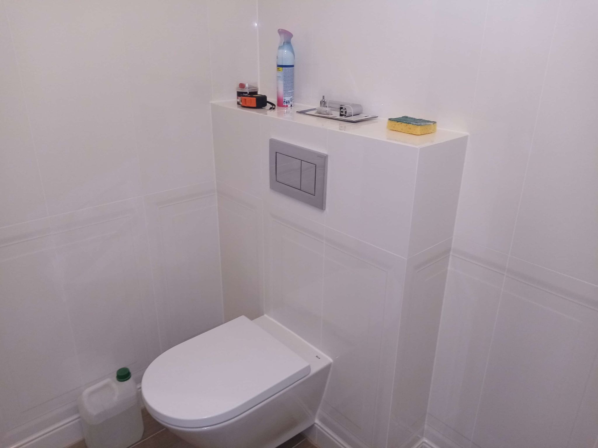 Nowoczesna, podwieszana toaleta w jasnej łazience z białymi płytkami. Na zabudowie stelaża widoczne akcesoria łazienkowe i przycisk spłukujący.