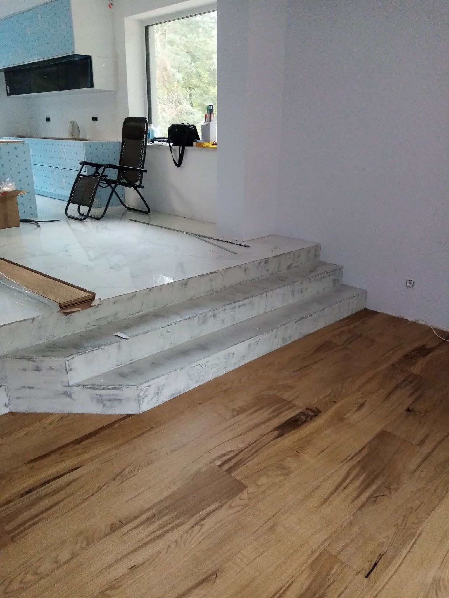 Wnętrze w trakcie wykańczania: drewniana podłoga ułożona, schody wyłożone płytkami imitującymi marmur, widoczne przygotowania do montażu kuchni.
