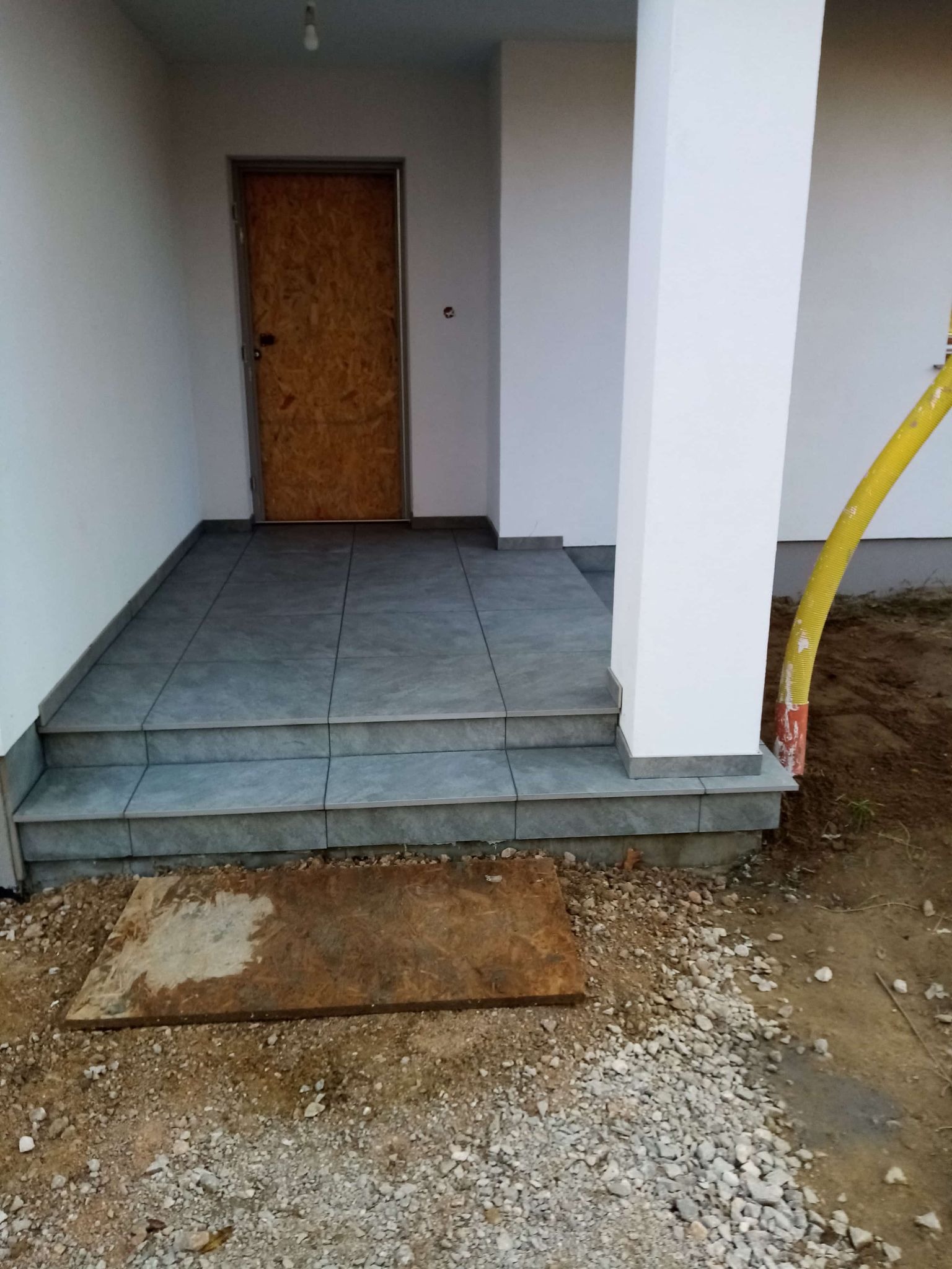 Wykończone płytkami schody wejściowe do domu z kolumną i żółtą rurą w tle. Drzwi zabezpieczone płytą. Widoczne niedokończone prace ziemne.