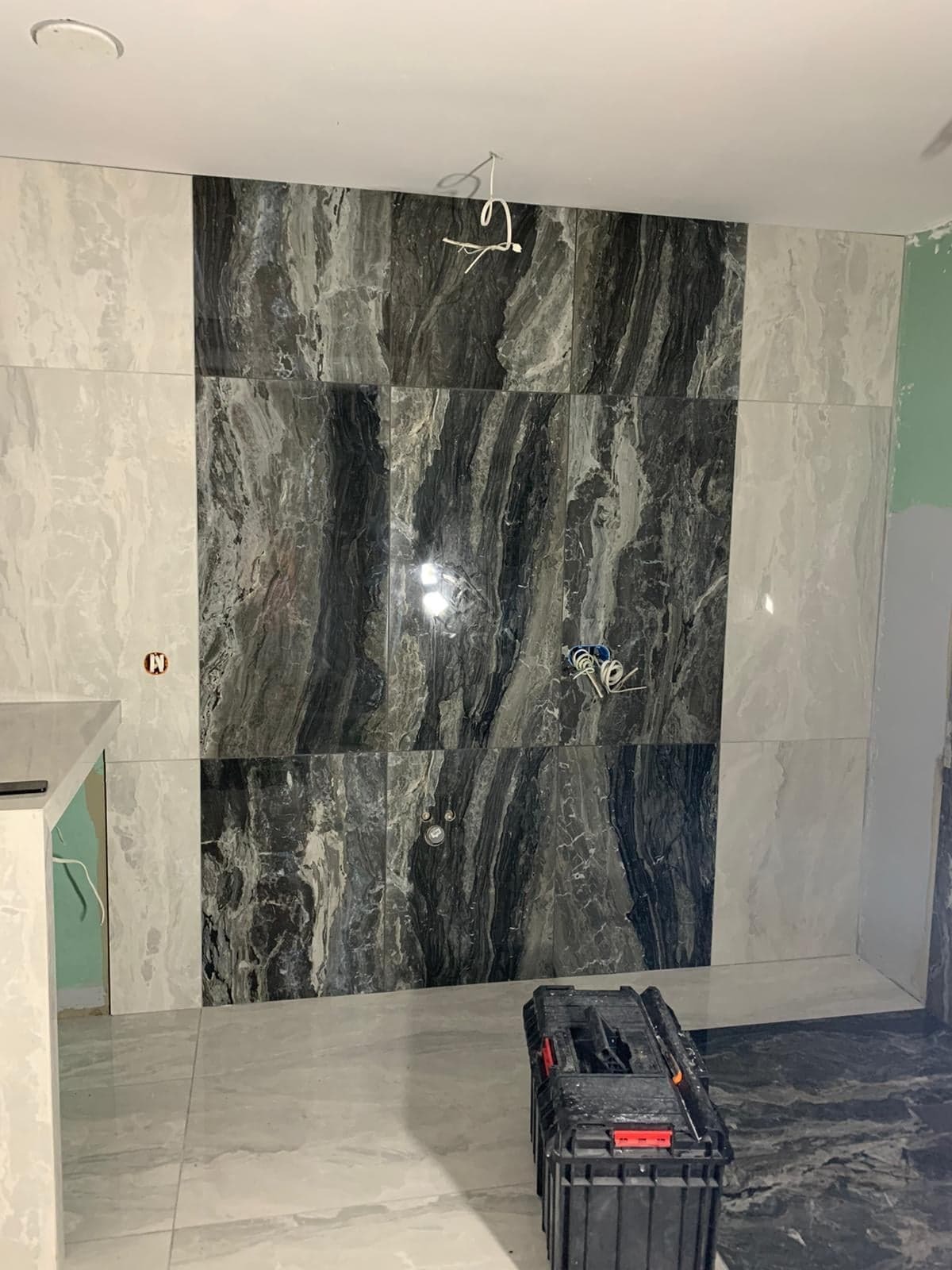 Wnętrze łazienki w trakcie prac. Ułożone płytki w odcieniach szarości i bieli, widoczna skrzynka z narzędziami i wystające kable.