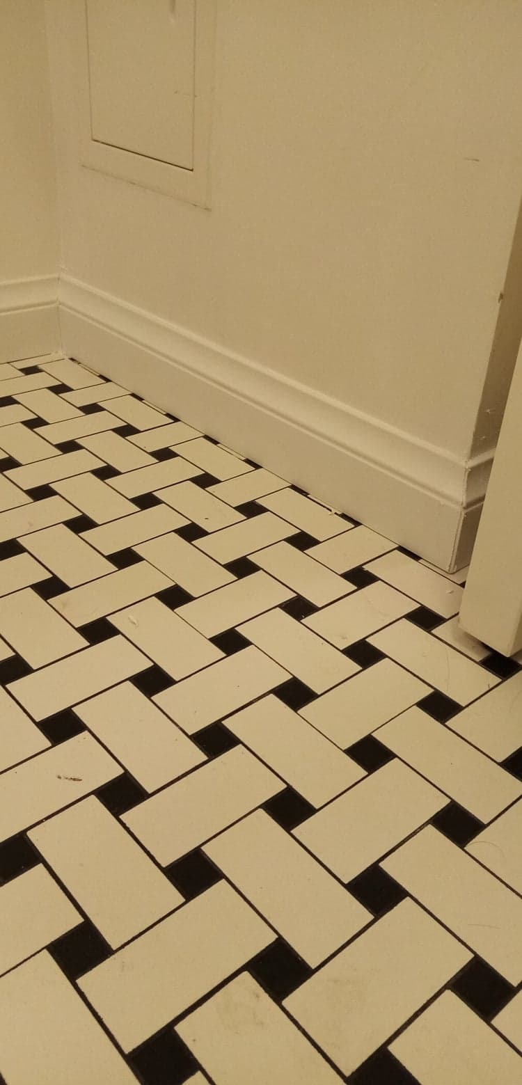 Ukończona podłoga z kremowych i czarnych płytek ułożonych w jodełkę, przy białej ścianie z listwą przypodłogową i fragmentem drzwi po prawej.