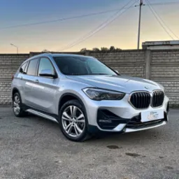 Srebrny SUV marki BMW X1, zaparkowany przed betonowym ogrodzeniem, w tle widoczne słupy energetyczne i budynki o zmierzchu.