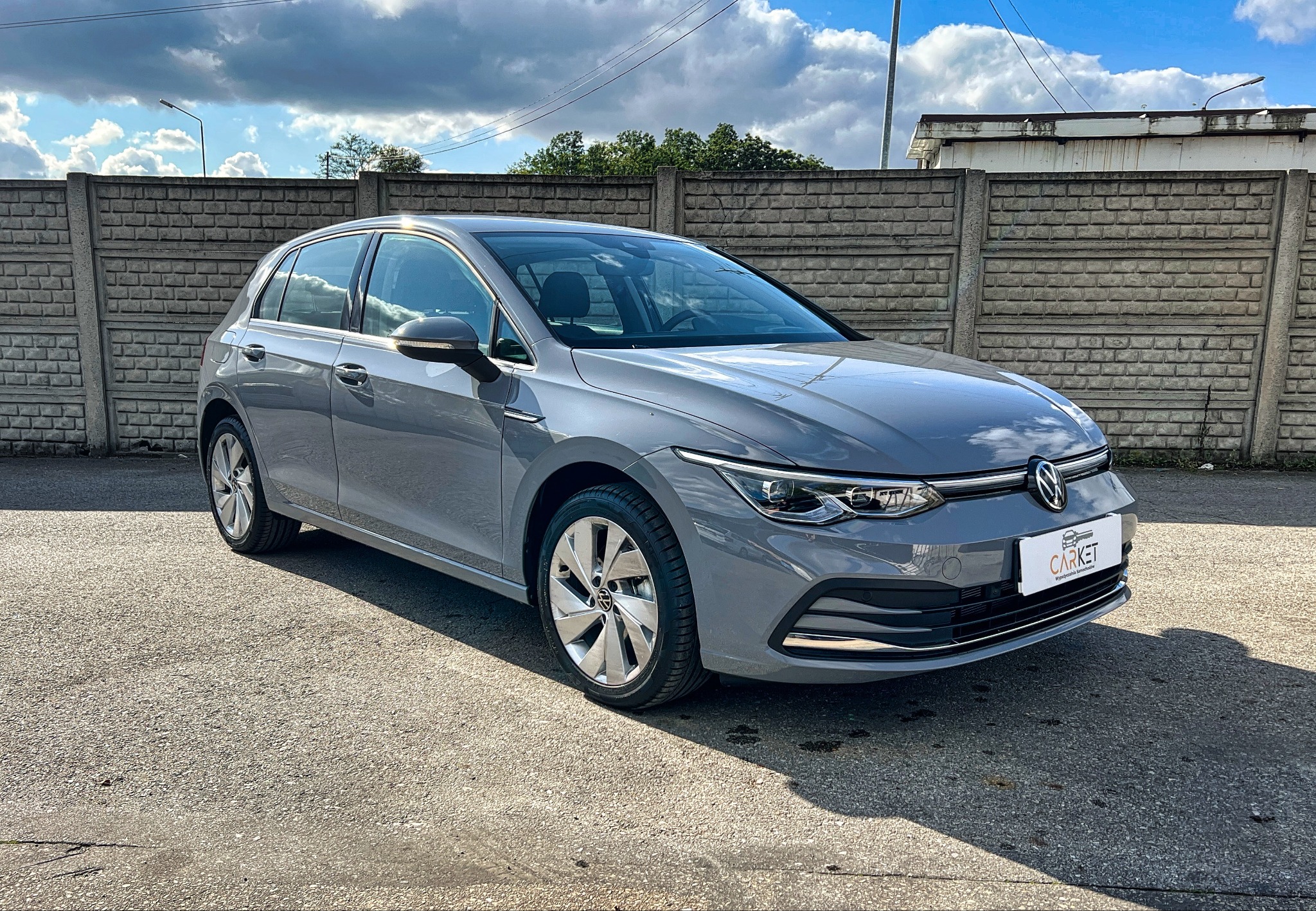 Szary Volkswagen Golf 8 generacji na tle betonowego muru i pochmurnego nieba, widoczny cień rzucany przez auto, logo firmy CARKET na przednim zderzaku.