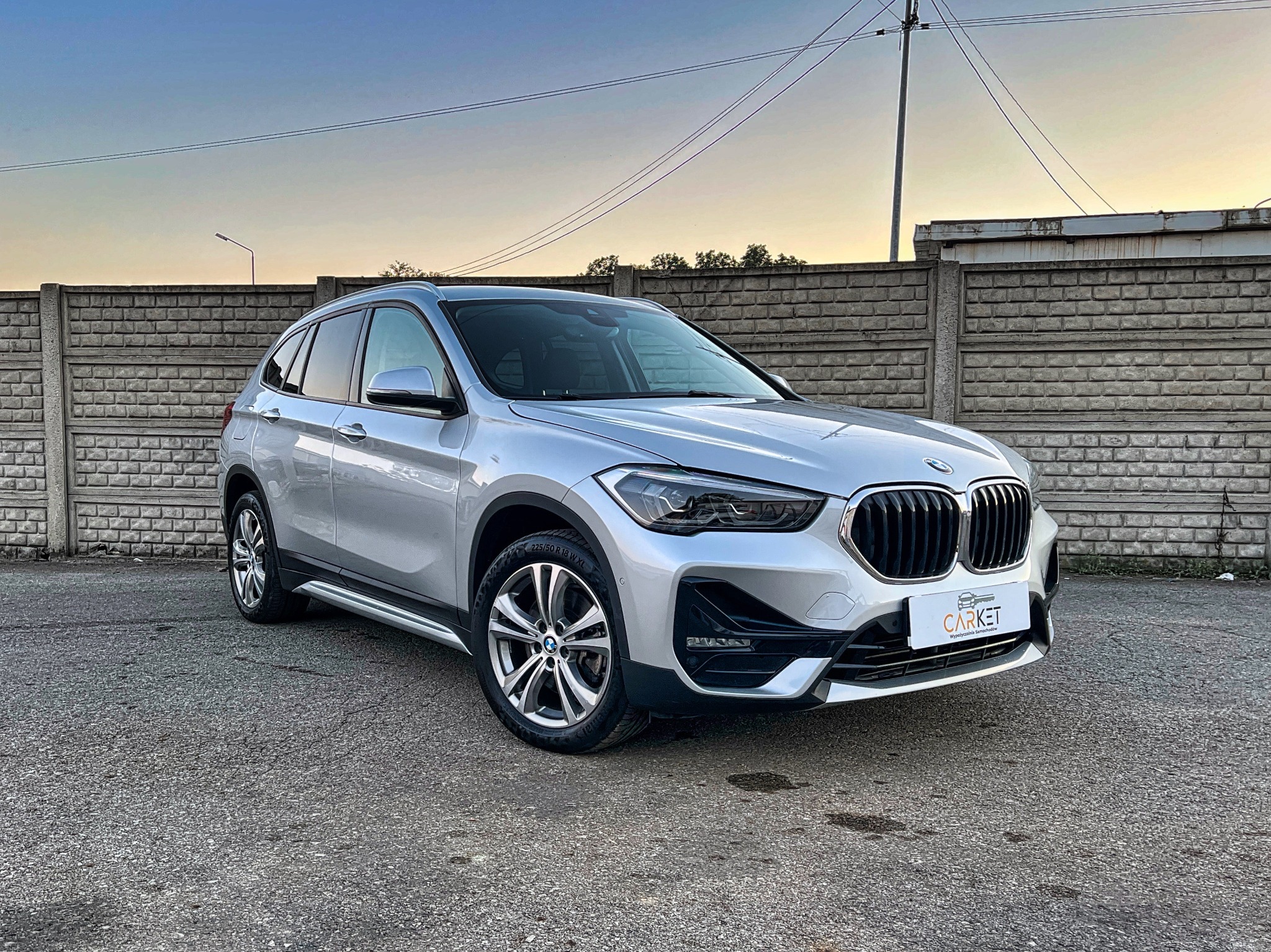 Srebrny SUV marki BMW X1, zaparkowany przed betonowym ogrodzeniem, w tle widoczne słupy energetyczne i budynki o zmierzchu.