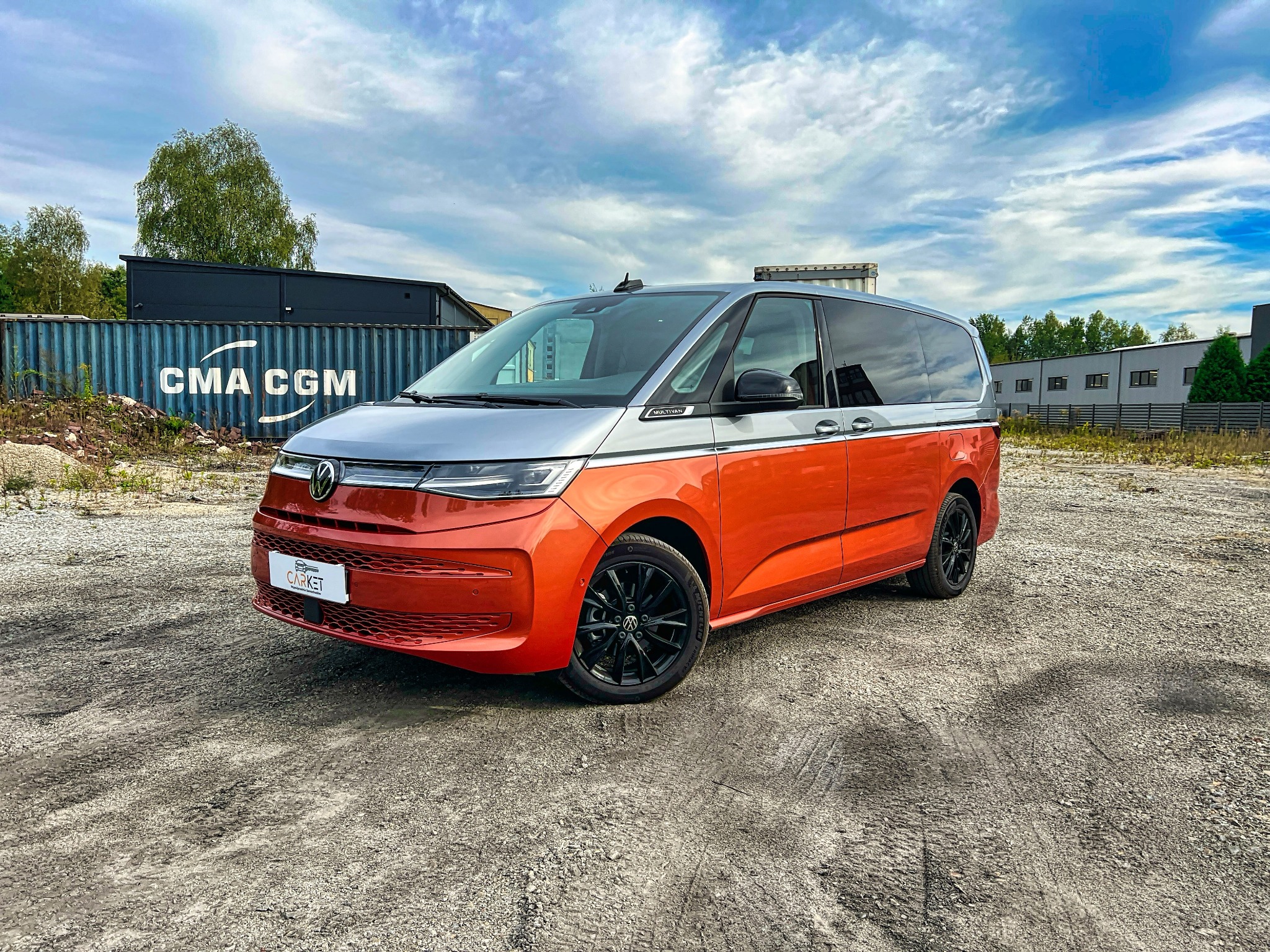 Pomarańczowo-srebrny van z czarnymi felgami na żwirowym placu, kontenerowiec w tle.