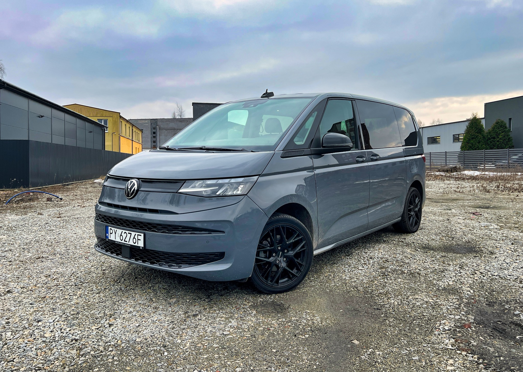 Szary Volkswagen Transporter T7 na żwirowym parkingu z czarnymi felgami, widok z przodu, pochmurne niebo w tle.