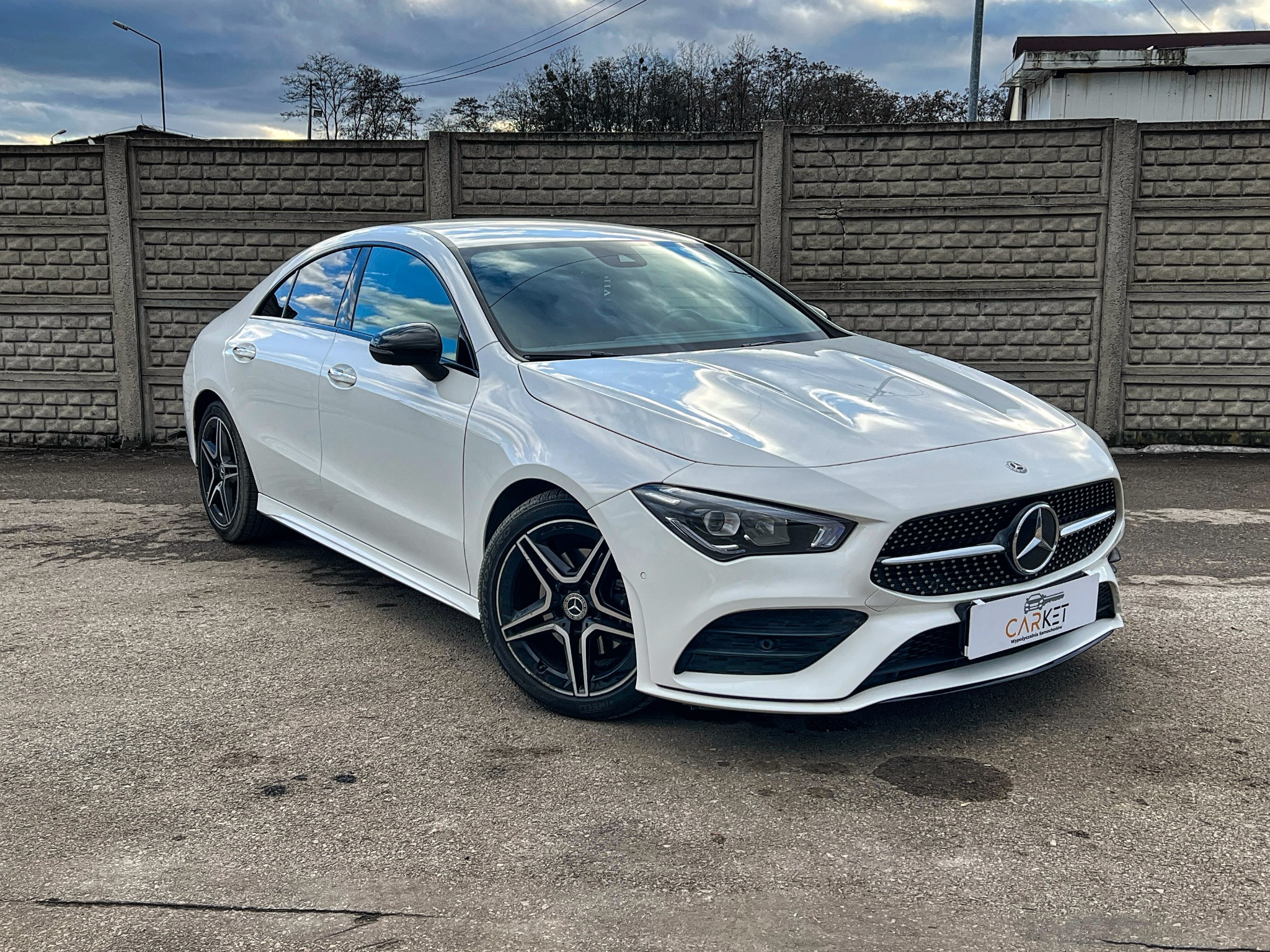 Biały samochód osobowy marki Mercedes-Benz CLA z czarnymi felgami, zaparkowany na szarym asfalcie na tle betonowego muru z widocznym logo firmy na tabliczce z przodu.