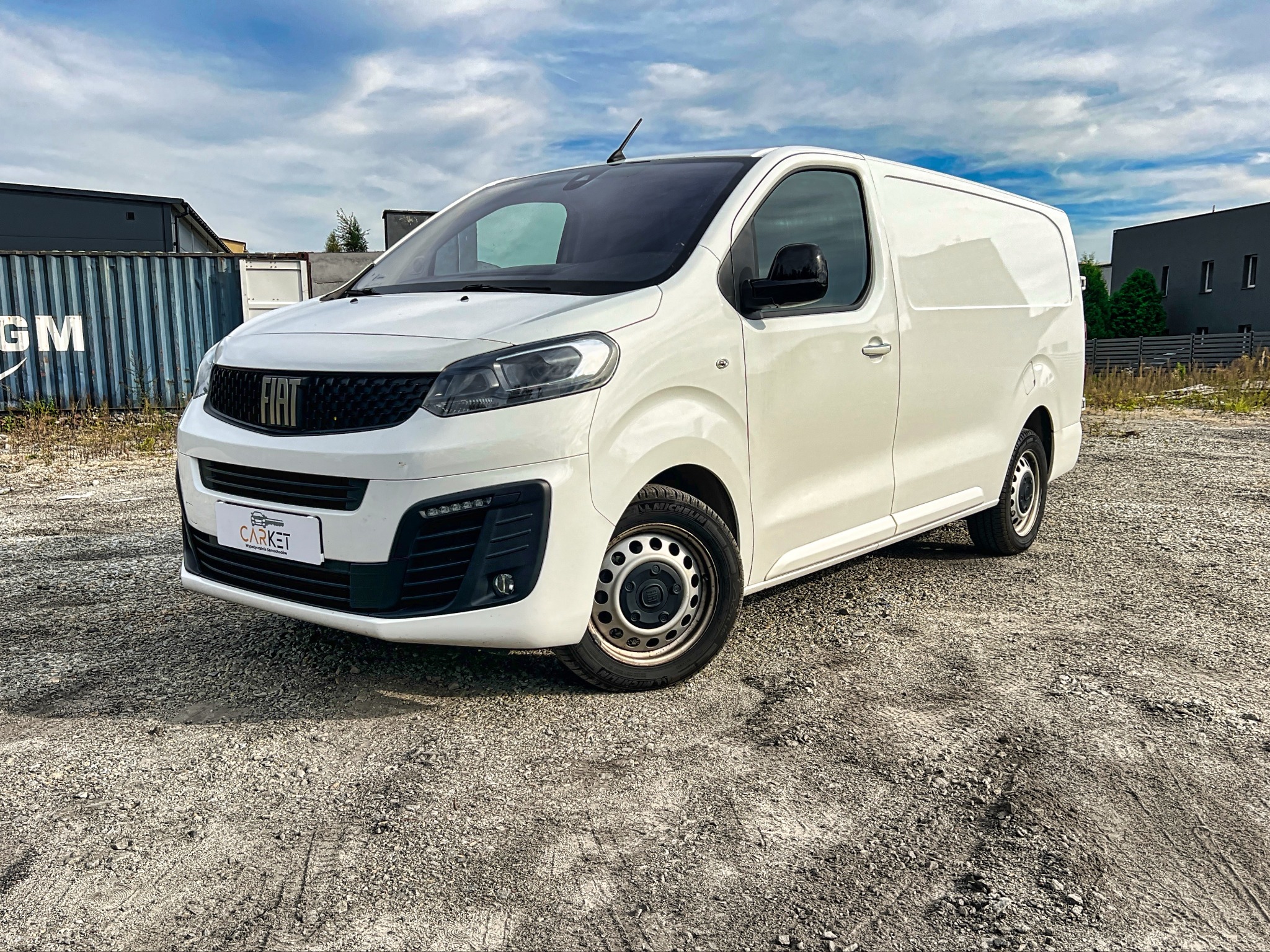 Biały samochód dostawczy Fiat, zaparkowany na żwirowym placu w Chrzanowie, z widocznym logo wypożyczalni na przednim zderzaku.